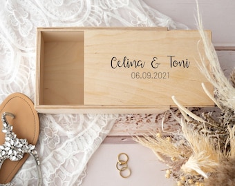 May include: A wooden box with a sliding lid, engraved with the names "Celina & Toni" and the date "06.09.2021". The box is surrounded by white lace, dried flowers, and two pairs of sandals.