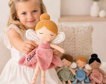 May include: A plush fairy doll with a mustard yellow corduroy bun, closed eyes, and a pink velvet dress featuring the embroidered name Celina. The doll has white wings and pink ballet-style leg ribbons. Three additional dolls are visible nearby.