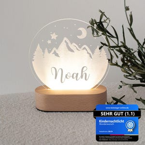 Peut inclure: Une veilleuse en acrylique blanche avec un motif de montagne et de lune. La lumière est allumée et le nom "Noah" est gravé sur le devant. La lumière repose sur une base en bois.