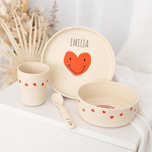 Könnte beinhalten: Ein beigefarbenes Kindergeschirr-Set mit einem roten Herzdesign. Der Teller trägt den Namen "Emilia". Das Set enthält eine Schüssel, einen Teller, eine Tasse und einen Löffel.