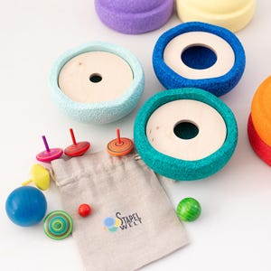 Può includere: Un giocattolo di impilamento in legno colorato con un arcobaleno di colori, tra cui viola, giallo, blu, verde, arancione e rosso. Il giocattolo viene fornito con un piccolo sacchetto di trottole e palline di legno.
