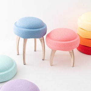 Könnte beinhalten: Fünf bunte, runde Hocker aus Schaumstoff mit Holzbeinen. Die Hocker sind in einem Halbkreis auf einer weißen Oberfläche angeordnet. Die Hocker sind mit "Tinu" und "Celina" beschriftet.