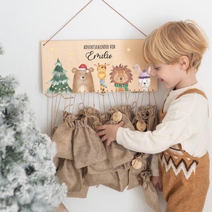 Può includere: Una targa rettangolare in legno con il testo "ADVENTSKALENDER VON Emilio" e illustrazioni ad acquerello di un albero di Natale, un orso con cappello da Babbo Natale, una giraffa, un leone e un coniglio. Ventiquattro piccoli sacchetti di juta marrone.