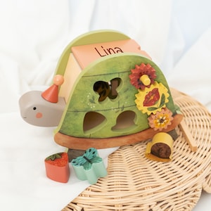 May include: A wooden turtle toy with a personalised name, "Lina", on the top. The turtle has a green shell with cut-out shapes and colourful flowers and butterflies. The toy comes with a set of colourful wooden shapes that fit into the cut-outs.