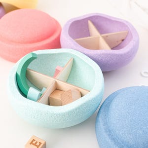 Può includere: Quattro contenitori rotondi con coperchio di colore pastello con una superficie testurizzata. I contenitori sono aperti e rivelano un divisorio in legno all'interno. Il contenitore al centro è blu chiaro e ha una forma di mezzaluna verde e un blocco di legno all'interno. Un blocco di legno con la lettera "E" è in primo piano.