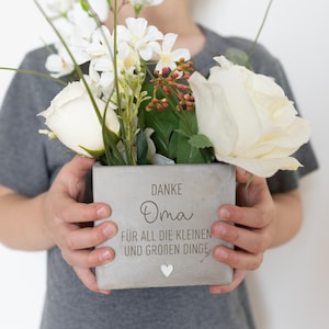 Könnte beinhalten: Ein grauer quadratischer Betonblumentopf gefüllt mit weißen Blumen und Grün. Der Topf trägt die Worte "DANKE Oma FÜR ALL DIE KLEINEN UND GROBEN DINGE" und ein kleines Herz. Der Topf wird gehalten.
