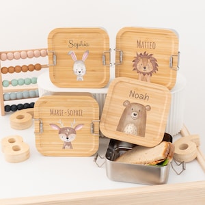 Op de afbeelding: Vier bamboe lunchboxen met dierenillustraties en namen: Sophia, Matteo, Marie-Sophie en Noah. Eén doos is open en onthult een roestvrijstalen container met een sandwich. Er zijn ook een telraam en houten cijfers aanwezig.