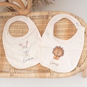 May include: Two off-white fabric baby bibs with snap closures. One bib features a watercolour illustration of a grey rabbit and the name Emma. The other bib features a watercolour illustration of a brown lion and the name Louis.