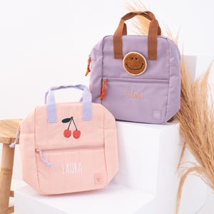 Op de afbeelding: Twee gewatteerde lunchtassen. Eén is lichtroze met een kersendesign en de naam "LAURA". De andere is lavendelkleurig met een smiley-patch en de naam "Cleo". Beide hebben handvatten en ritszakken.