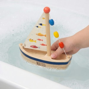 Könnte beinhalten: Ein hölzernes Spielzeug-Segelboot mit einem weißen Segel, das mit bunten Fischen verziert ist. Das Boot hat einen blauen Streifen und den Namen "MATTEO" auf der Seite aufgedruckt. Eine Schnur mit bunten Bojen hängt am Mast. Das Boot befindet sich im Wasser.