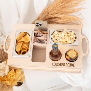 Può includere: Vassoio di servizio in legno con scomparti per snack e bevande. Ha un supporto per telefono integrato, uno scomparto per patatine, uno scomparto per popcorn, uno scomparto per snack e due portabevande. Il vassoio è etichettato "Couchbar Deluxe".