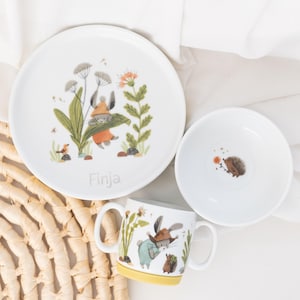 Pode incluir: Um conjunto de louça de cerâmica branca para crianças com tema de floresta. O conjunto inclui um prato, uma tigela e uma xícara. O prato apresenta um coelho segurando uma folha, a tigela apresenta um ouriço e a xícara apresenta um coelho e um ouriço. O prato tem o nome "Finja" escrito nele.