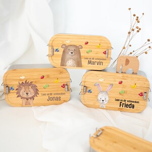 Peut inclure: Trois boîtes à lunch en bois avec des illustrations d'animaux et des noms personnalisés. Les boîtes à lunch sont brunes avec un motif de grain de bois clair. La première boîte à lunch présente un ours avec le nom "Marvin". La deuxième boîte à lunch présente un lion avec le nom "Jonas". La troisième boîte à lunch présente un lapin avec le nom "Frieda".