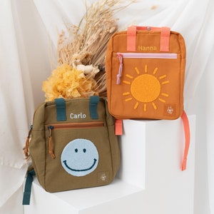 Może przedstawiać: Dwa małe plecaki z imionami. Jeden plecak jest brązowy z wyszytym żółtym słońcem z przodu, a drugi jest zielony z wyszytą niebieską uśmiechniętą buźką z przodu.
