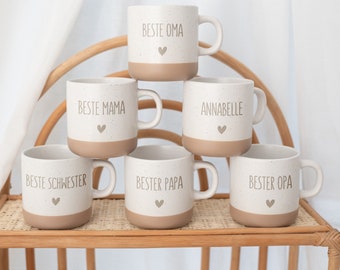 May include: A set of six ceramic mugs with a speckled cream upper section and a solid tan base. Each mug features a small heart icon and text including BESTE OMA, BESTE MAMA, ANNABELLE, BESTE SCHWESTER, BESTER PAPA, and BESTER OPA in a simple font.