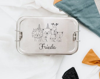 Peut inclure: Une boîte à lunch en argent avec une illustration en noir et blanc d'animaux et le nom "Frieda" imprimé sur le devant. La boîte à lunch est sur une surface blanche avec un triangle en tissu multicolore et deux nœuds papillon.