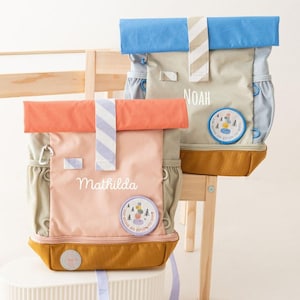 Op de afbeelding: Twee kleurrijke rugzakken met gepersonaliseerde namen en patches. De ene rugzak is perzik en oranje, de andere is blauw en bruin. Beide hebben gestreepte accenten en een ronde patch met een natuurscène. De rugzakken staan op een wit oppervlak.