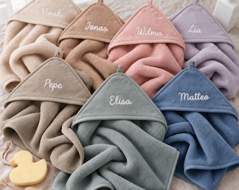May include: A collection of six hooded towels in beige, tan, pink, lavender, gray, and blue, each embroidered with a name: Noah, Jonas, Wilma, Lia, Pepe, and Elisa. A yellow rubber duck sits near the towels, which are made of soft terry cloth.