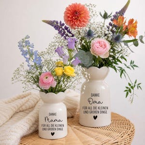 May include: Two white ceramic vases with scalloped rims, each featuring black text. The smaller vase reads "DANKE Mama FÜR ALL DIE KLEINEN UND GROBEN DINGE" with a heart, and the larger reads "DANKE Oma FÜR ALL DIE KLEINEN UND GROBEN DINGE" with a heart.