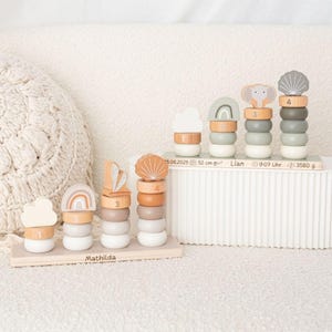 Op de afbeelding: Houten stapelspeelgoed in pastelkleuren met wolken, regenboog- en schelpvormen. Het speelgoed is gerangschikt op een wit oppervlak, met de naam "Mathilda" en baby-informatie. De lengte van de baby is 52 cm.