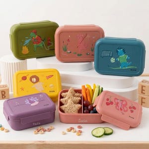 Könnte beinhalten: Eine Sammlung bunter Brotdosen mit Cartoon-Tierdesigns. Die Dosen sind in verschiedenen Farben erhältlich, darunter Grün, Rosa, Blau, Gelb und Lila. Eine Dose ist geöffnet und zeigt ein Lunchpaket mit Sandwiches, Gemüse und Obst. Die Marke "trixie" ist sichtbar.