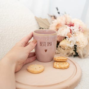 Puede incluir: Una taza de cer&aacute;mica rosa con el texto "BESTE OMA" y un s&iacute;mbolo de coraz&oacute;n. La taza est&aacute; sostenida por una mano y se encuentra sobre una bandeja de madera con galletas. Un ramo de flores est&aacute; en el fondo.