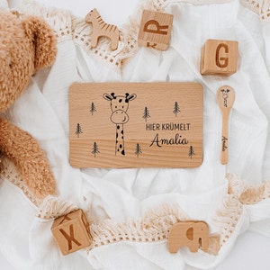 May include: A wooden plate with a giraffe illustration and the text "Hier kr&uuml;melt Amalia" in black. The plate is surrounded by wooden toys, including a giraffe, elephant, and blocks.