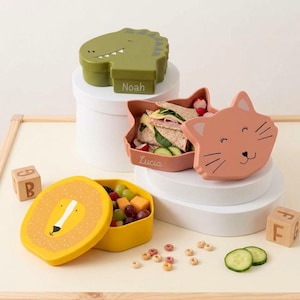 Könnte beinhalten: Drei tierförmige Brotdosen: ein grüner Dinosaurier, eine pfirsichfarbene Katze und ein gelber Löwe. Der Dinosaurier und die Katze sind geöffnet und zeigen Sandwiches und Snacks. Der Löwe enthält Obst. Die Dosen stehen auf einer weißen Oberfläche.