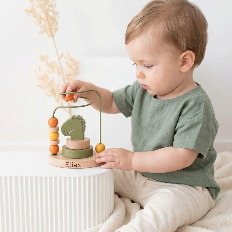 Peut inclure: Un jouet labyrinthe de perles en bois avec une t&ecirc;te de dinosaure verte, des perles orange et jaunes, et le nom "Elias" grav&eacute; sur la base. Le jouet a un cadre m&eacute;tallique pour faire glisser les perles. Con&ccedil;u pour les tout-petits.