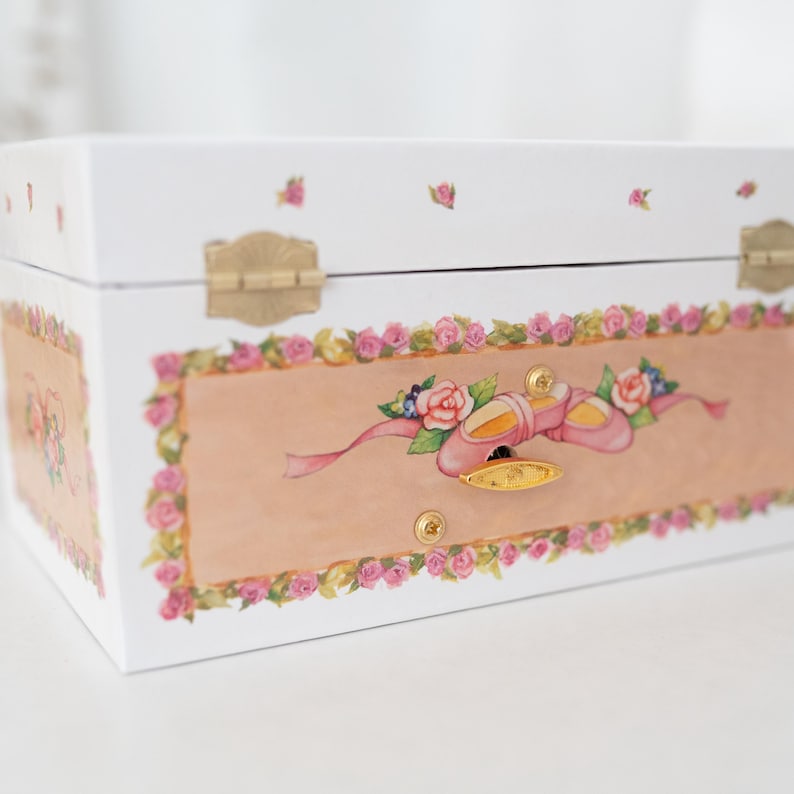 May include: White wooden music box with a pink and gold ballerina shoe design. The box has a floral border and a gold latch.