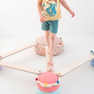 Könnte beinhalten: Ein Balancierbalken-Set bestehend aus hellen Holzplanken, die mit runden, gepolsterten Plattformen verbunden sind. Die Plattformen sind in verschiedenen Farben wie Rosa, Hellblau und einem mehrfarbigen Sprenkelmuster gehalten und mit kleinen farbigen Stiften befestigt.