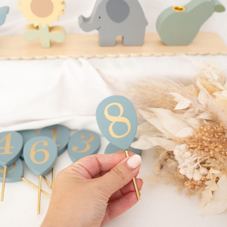 May include: A hand holding a light blue wooden balloon-shaped number eight with a gold number. Other wooden numbers and animal figures are in the background. The balloon is on a gold stick.