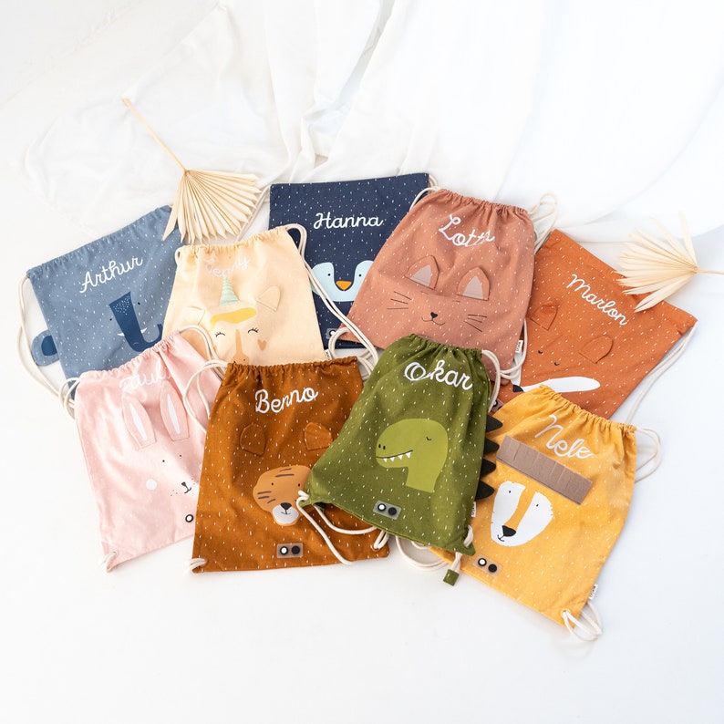 May include: Nine colorful drawstring bags with various animal designs and personalized names, including Arthur, Hanna, Lotti, Marlon, Benno, Oskar, Nole. The bags are made of a soft, textured fabric and have a drawstring closure.