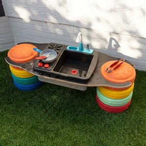 Könnte beinhalten: Ein Spielküchen-Set aus Kunststoff mit schwarzer Arbeitsplatte, Spülbecken, Wasserhahn und Herdplatten. Die Basis besteht aus Stapeln bunter orangefarbener, gelber, türkisfarbener und roter Scheiben. Enthält eine kleine Pfanne und eine Zange.
