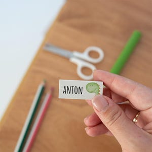 May include: A rectangular white eraser featuring the name ANTON printed in black capital letters next to a small green cartoon dinosaur illustration. The eraser is held against a blurred background containing pencils and scissors.