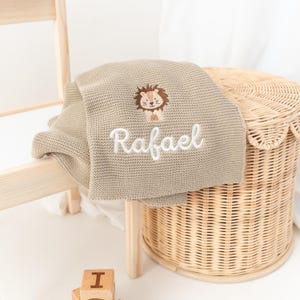 Puede incluir: Una manta de beb&eacute; de punto beige con el nombre "Rafael" bordado en escritura blanca. La manta presenta un dise&ntilde;o de le&oacute;n bordado. La manta est&aacute; sobre una silla de madera y una cesta de mimbre.