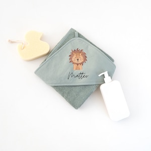 May include: A hooded towel with a lion design and the name Matteo embroidered on it. The towel is green and has a soft texture. A yellow sponge and a white bottle of lotion are also in the image.