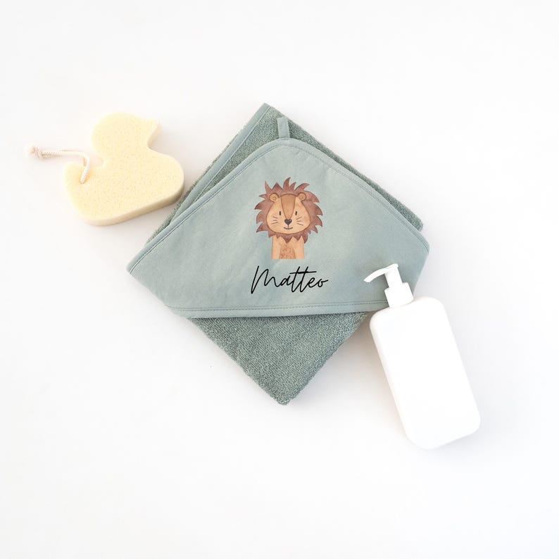 May include: A hooded towel with a lion design and the name Matteo embroidered on it. The towel is green and has a soft texture. A yellow sponge and a white bottle of lotion are also in the image.