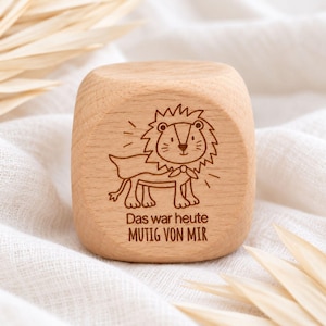 May include: A light-coloured wooden cube featuring a laser-engraved illustration of a lion wearing a cape. Below the lion, the text reads: Das war heute MUTIG VON MIR. The cube has rounded edges and a smooth, natural wood grain finish throughout.