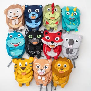 Peut inclure: Une collection de sacs à dos colorés sur le thème des animaux. Les modèles comprennent un singe, un ours, un dinosaure, un tricératops, un requin, un chat, un renard, un koala, un tigre, un chat et un lion. Chaque sac à dos présente un visage d'animal unique et une étiquette nominative.