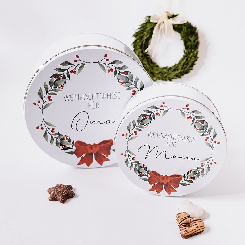 May include: Two white, round tins with lids, decorated with a wreath of green leaves and red berries. The tins have a red bow and the text "WEIHNACHTSKEKSE F&Uuml;R Oma" and "WEIHNACHTSKEKSE F&Uuml;R Mama". A small wreath and cookies are also present.