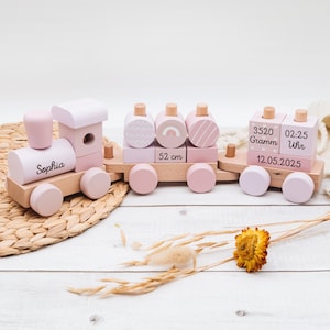 May include: A wooden toy train in pastel pink and natural wood tones. The train features the name "Sophia" on the engine and decorative blocks with patterns and measurements. The train is approximately 20.5 inches long.