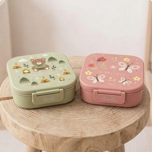 Peut inclure: Deux boîtes à lunch carrées. L'une est vert clair avec un ours et une scène de camping, l'autre est rose avec des papillons et des fleurs. Le nom "Noah" est sur la boîte verte et "Amalia" sur la boîte rose.