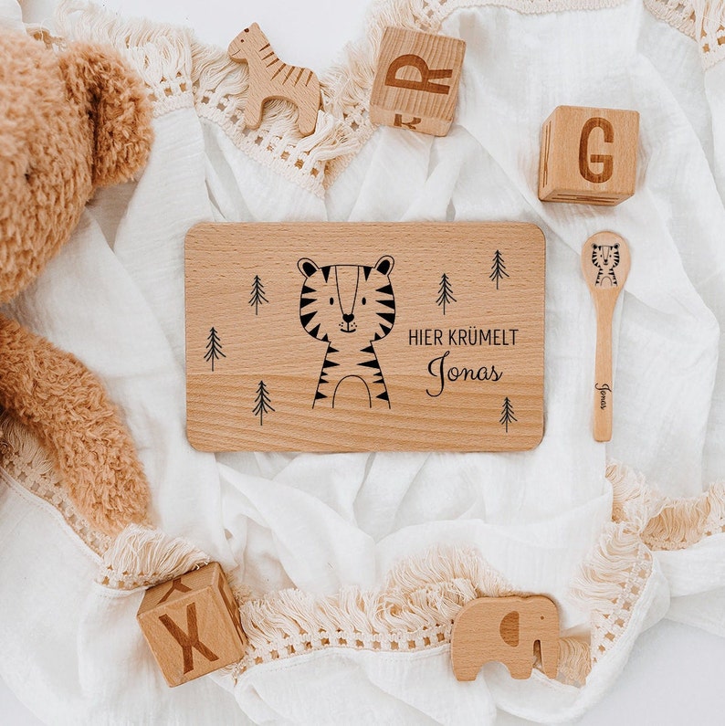 May include: Wooden tray with a tiger illustration and the text "Hier kr&uuml;melt Jonas". The tray is surrounded by wooden toys, including a spoon, a giraffe, an elephant, and blocks.