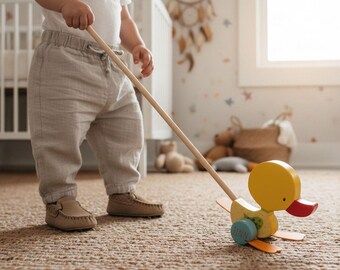 May include: A yellow wooden pull-along duck toy with orange wings and a red beak. The duck has blue wheels and is attached to a long wooden handle. The toy is being pulled across a woven rug.
