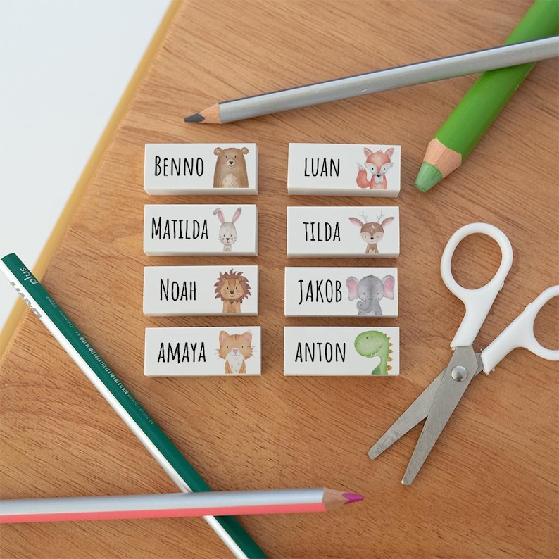 May include: Eight rectangular white labels featuring black printed names and watercolour animal illustrations including a bear, fox, rabbit, deer, lion, elephant, cat, and dinosaur, arranged with two pencils and a pair of white scissors.