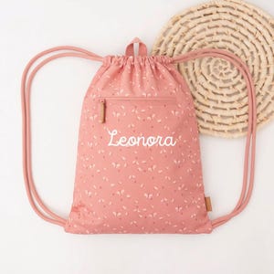 May include: A pink drawstring backpack with a floral pattern and the name "Leonora" embroidered in white. The bag has a zippered pocket and pink drawstrings. A woven circular placemat is in the background.