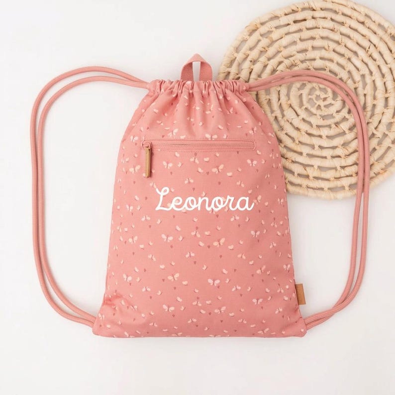 May include: A pink drawstring backpack with a floral pattern and the name "Leonora" embroidered in white. The bag has a zippered pocket and pink drawstrings. A woven circular placemat is in the background.