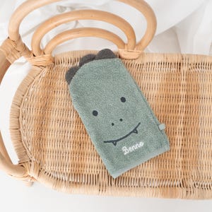 May include: A green dinosaur-shaped washcloth with a smiling face and the name "Benno" embroidered on it. The washcloth is lying on a woven wicker chair.