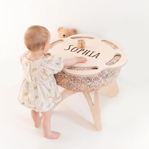 Op de afbeelding: Een ronde houten tafel met een bloemvormig blad waarop de naam SOPHIA staat en een klein houten blokje met de letter T. De tafel heeft een basis bekleed met kleurrijke confetti-stof en staat op poten van lichtgekleurd hout.